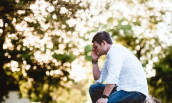 Mann mit Hemd sitzt in der Natur und verdeckt Augen mit der Hand als Zeichen von Stress